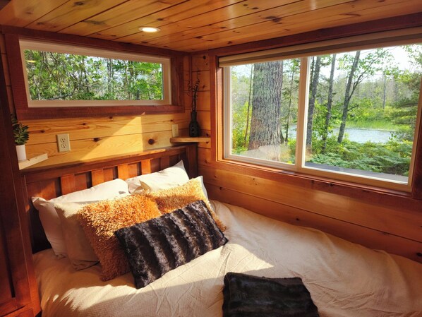 A wooden cabin bedroom with a bed, pillows, a nightstand, a window with a view of a forest and a lake.