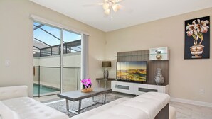 A modern living room with a sofa, a coffee table, and a TV. There's a sliding glass door leading to a balcony. - Four Bedroom Close to Disney w Pool 4898 (Kissimmee)