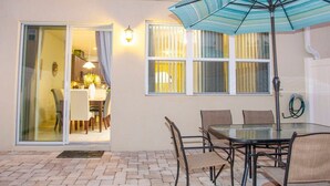 Um pátio externo com mesa e cadeiras de vidro, um guarda-sol azul e uma porta de correr de vidro que dá para uma área de refeições interna.