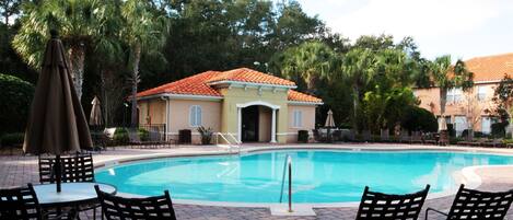 A swimming pool with lounge chairs and umbrellas, surrounded by palm trees and a building with a tiled roof.