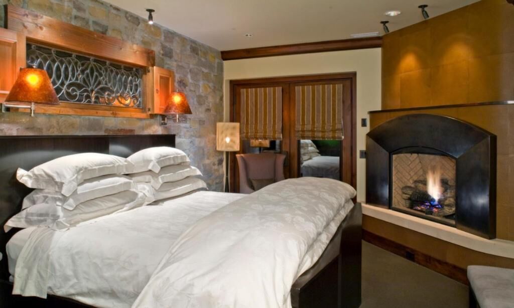 A bedroom with a stone accent wall, a fireplace, a bed with white linens, and a nightstand with a lamp.