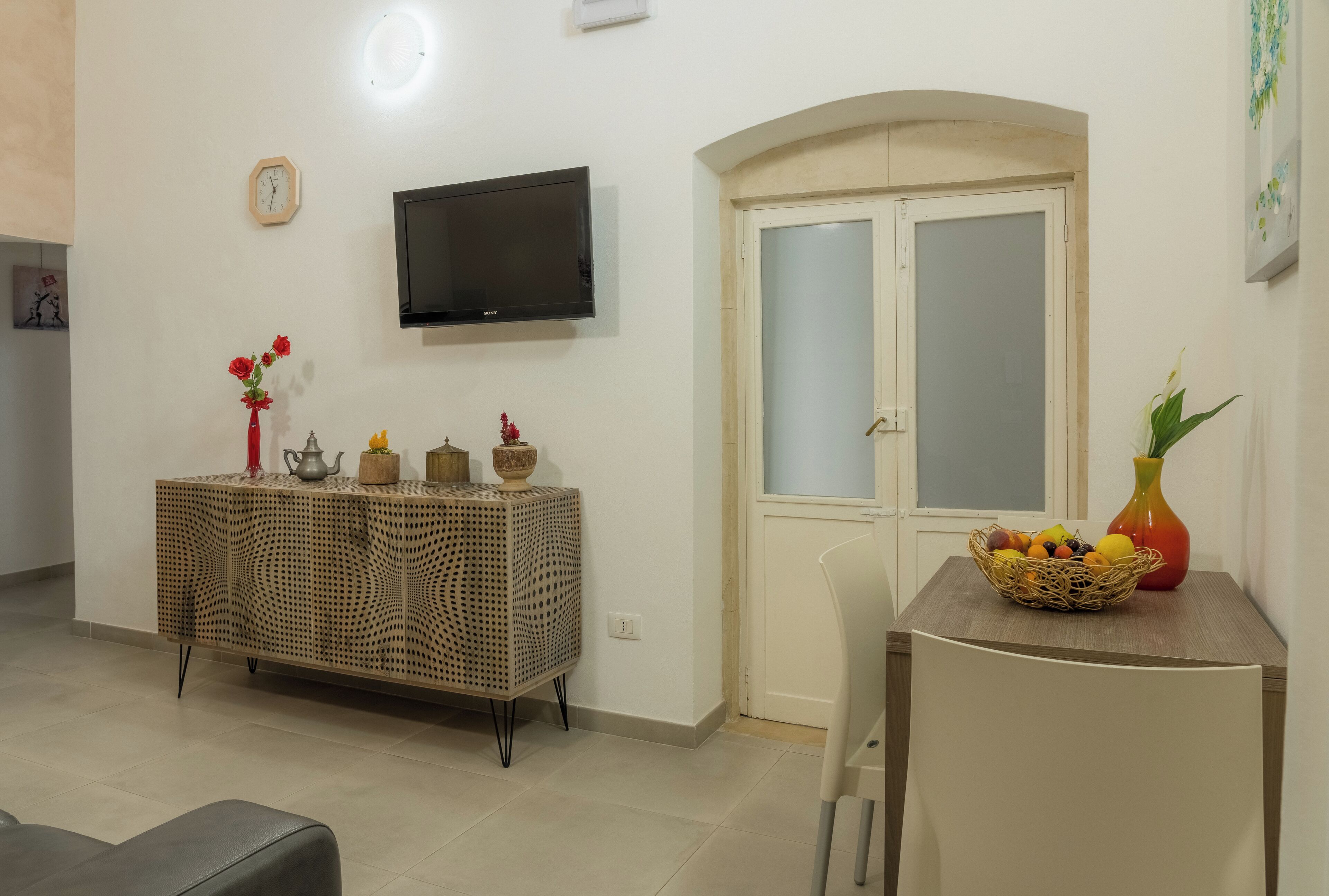 A living room with a television, a decorative cabinet, a door, and a table with a fruit basket.