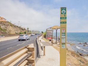 A coastal road with a bus stop shelter, a signpost indicating bus routes, and a view of the sea.