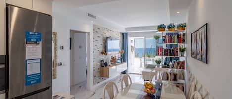 Une cuisine moderne avec un réfrigérateur en acier inoxydable, un coin repas doté d’une table en verre et une vue sur la mer par la porte ouverte.