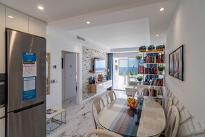 Une cuisine moderne avec un réfrigérateur en acier inoxydable, un coin repas doté d’une table en verre et une vue sur la mer par la porte ouverte.