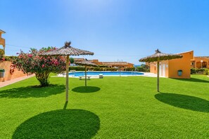 A swimming pool with a thatched umbrella, a well-maintained lawn, and a building with a tiled roof in the background.