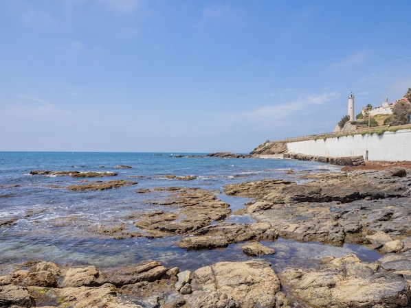 A rocky shoreline with clear water, a white wall, and a lighthouse in the background.
