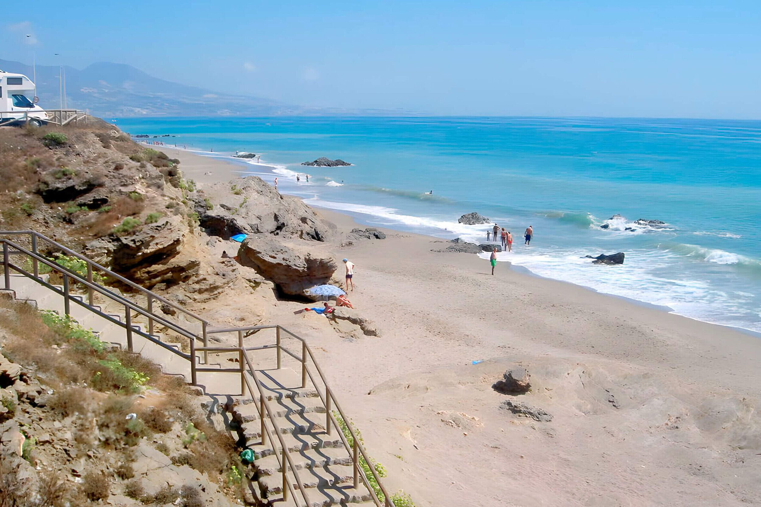 A beach with a staircase leading down to the shore, rocky outcrops, and clear blue water.