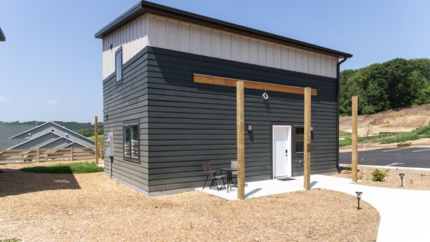 Een modern gebouw met een donkere gevel, een witte deur en een klein terras met twee stoelen.