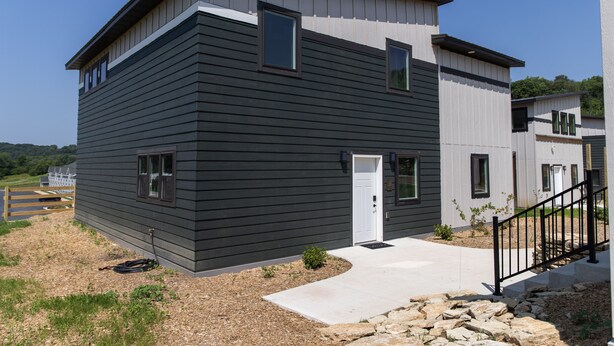 Une maison moderne à deux étages, dotée d’une façade sombre, d’une porte blanche et d’un petit jardin devant, avec un chemin menant à l’entrée.