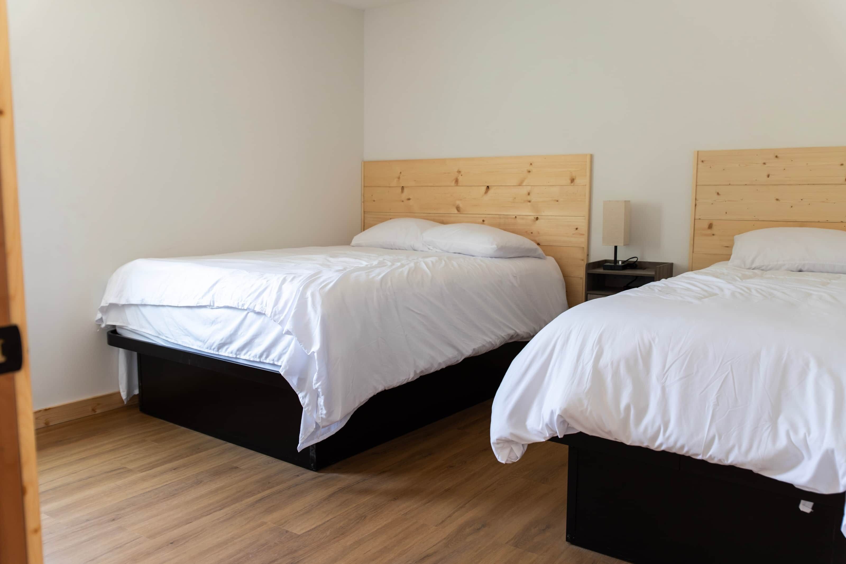 Ein Zimmer mit zwei Betten, jeweils mit einem Holz-Kopfteil, weißer Bettwäsche und einem Nachttisch mit Lampe.