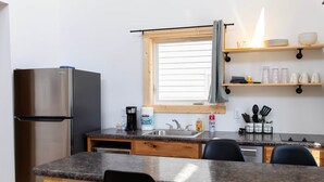 A compact kitchen with a stainless steel refrigerator, a sink, and a window with blinds. There are shelves above the sink holding dishes and glasses.