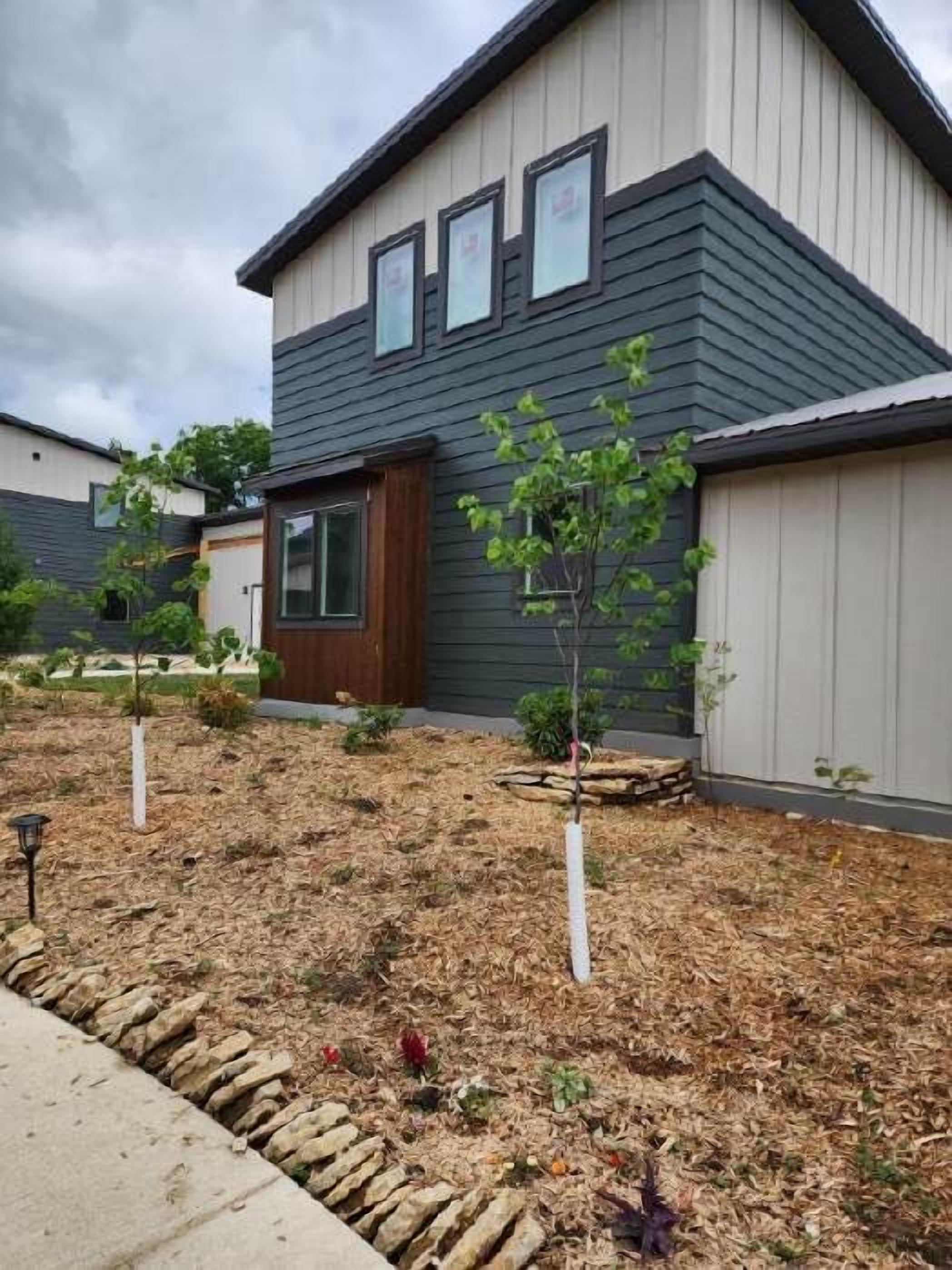 A modern two-story house with a dark grey exterior and white trim. There are three windows on the upper floor and a small front porch. The yard is landscaped with mulch and small plants.