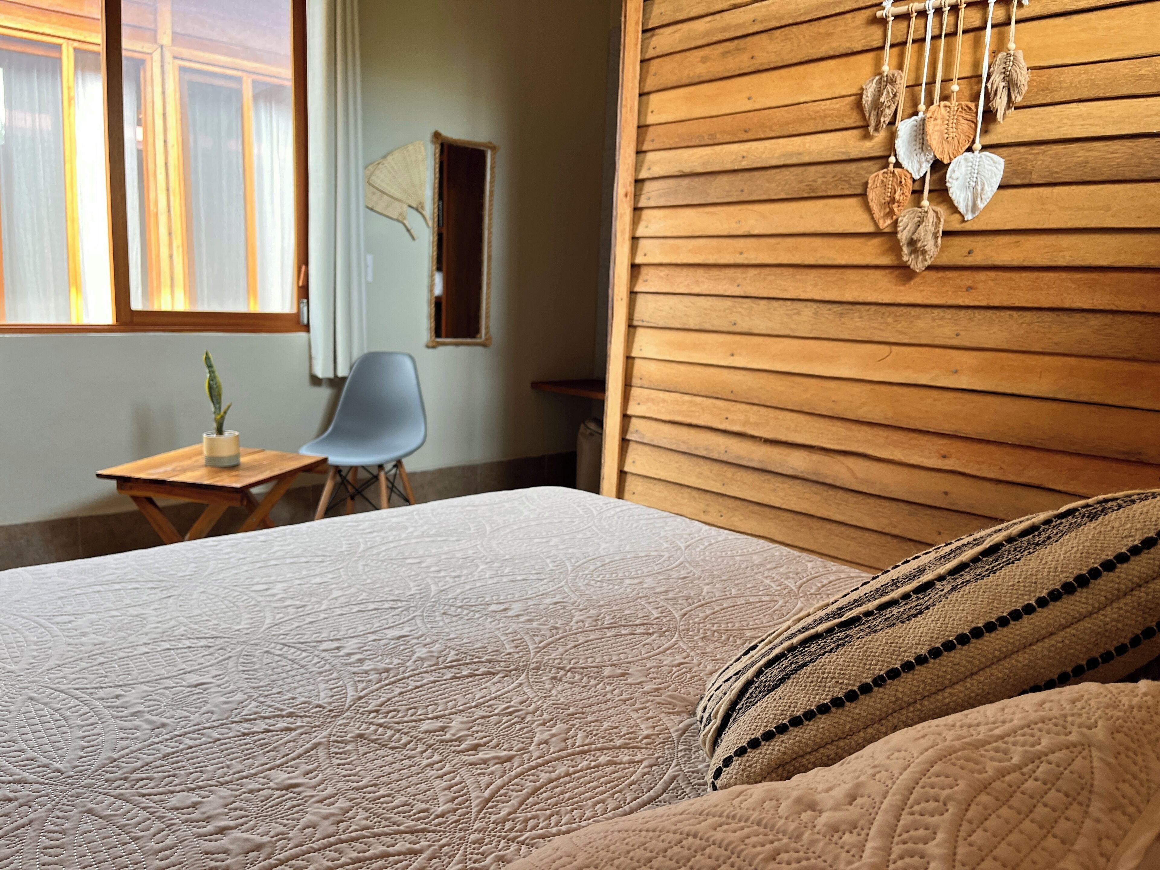 A bedroom with a wooden headboard, a bed with patterned bedding, a small table with a plant, a chair, and a wall-mounted decorative item.