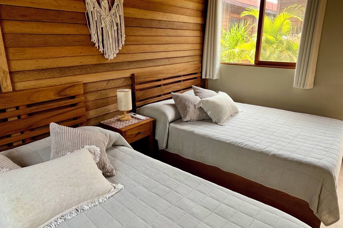 A room with two beds, wooden paneling, a macrame wall hanging, a nightstand with a lamp, and a window with a view of greenery.