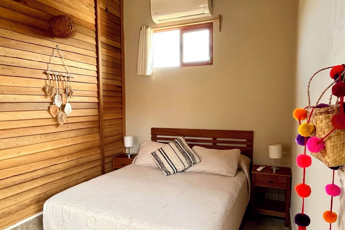 A bedroom with a wooden slat wall, a bed with a white bedspread, two bedside lamps, a window with blinds, and a wall-mounted decorative basket.