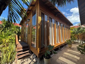 Una cabaña de madera con ventanas grandes, una terraza de madera y plantas tropicales alrededor.