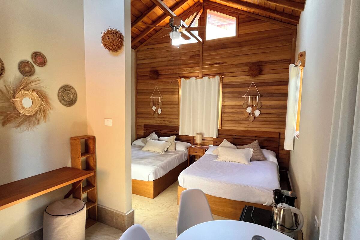 A cozy room with two beds, a small table, a bench, and a ceiling fan.