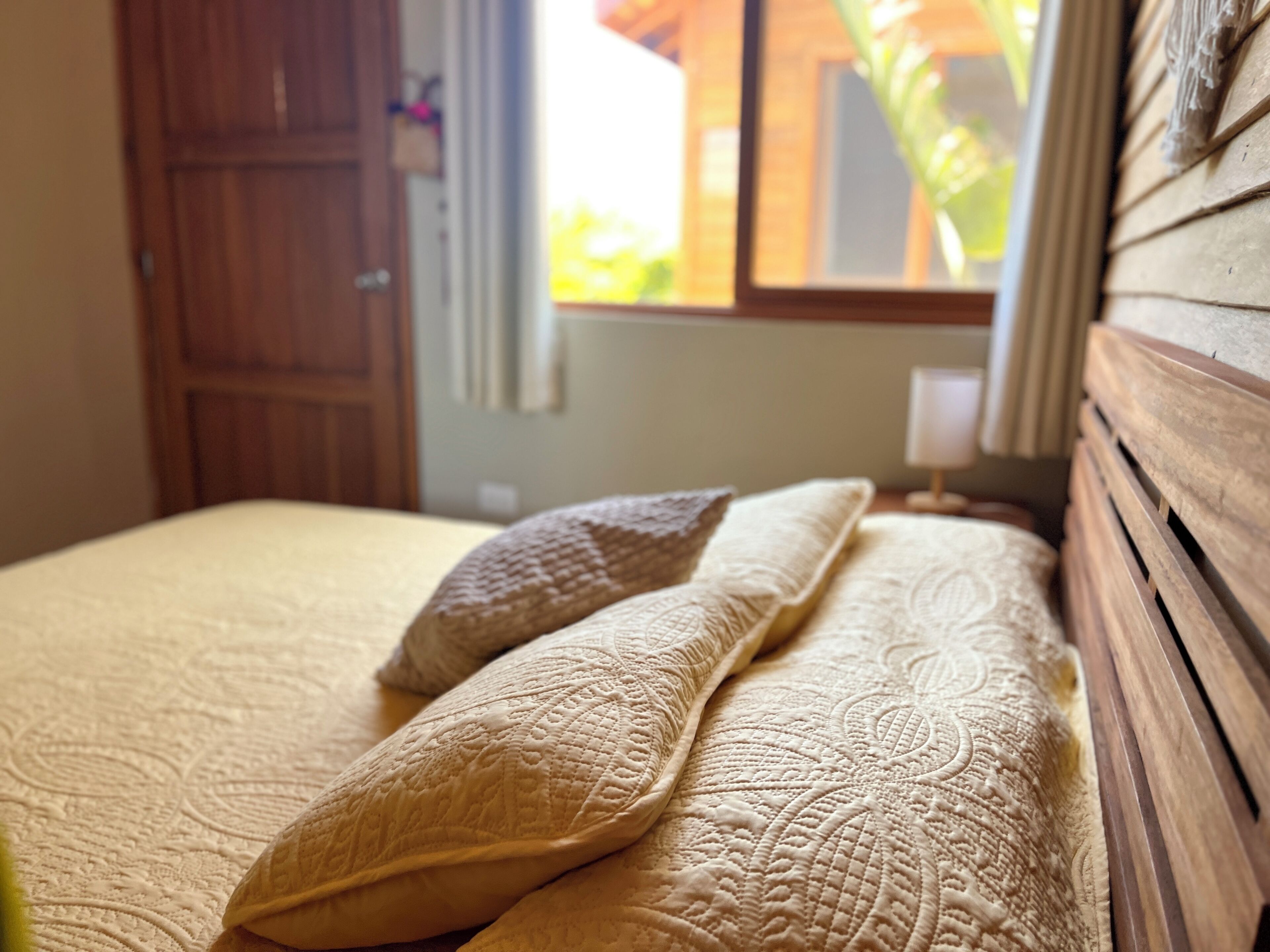 A bed with a patterned bedspread and two pillows, a wooden headboard, and a window with a view of greenery.