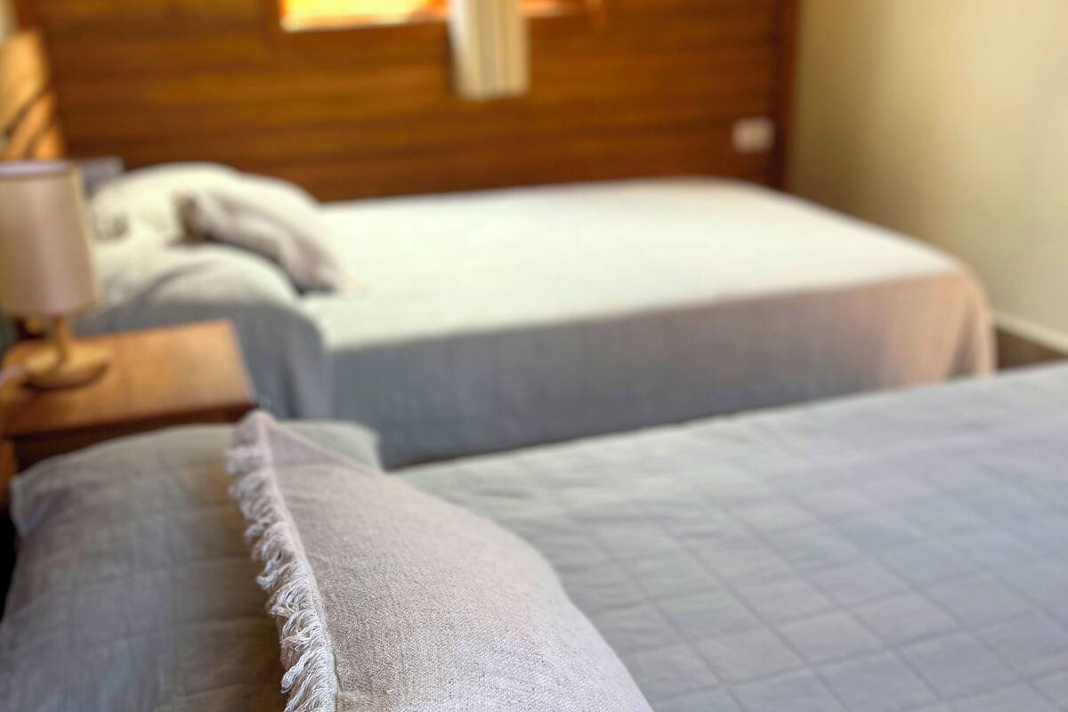 Two beds with headboards, a bedside table with a lamp, and a window with daylight.