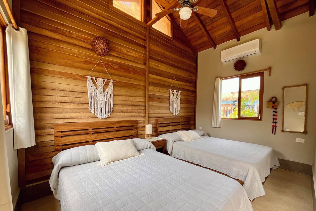 A room with wooden walls, two beds with white bedding, a ceiling fan, a window with a view, and a wall-mounted air conditioner.