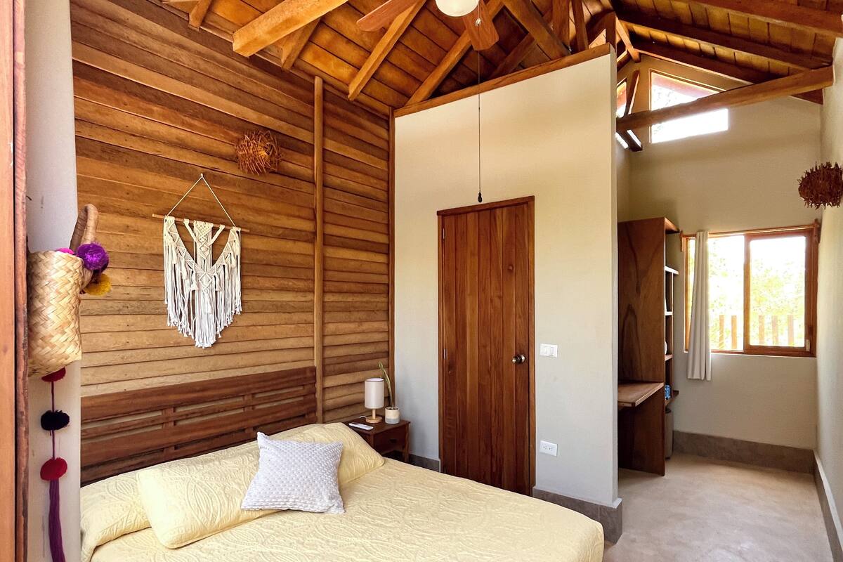A bedroom with a wooden ceiling, a bed with a white pillow, a macrame wall hanging, and a wooden door.