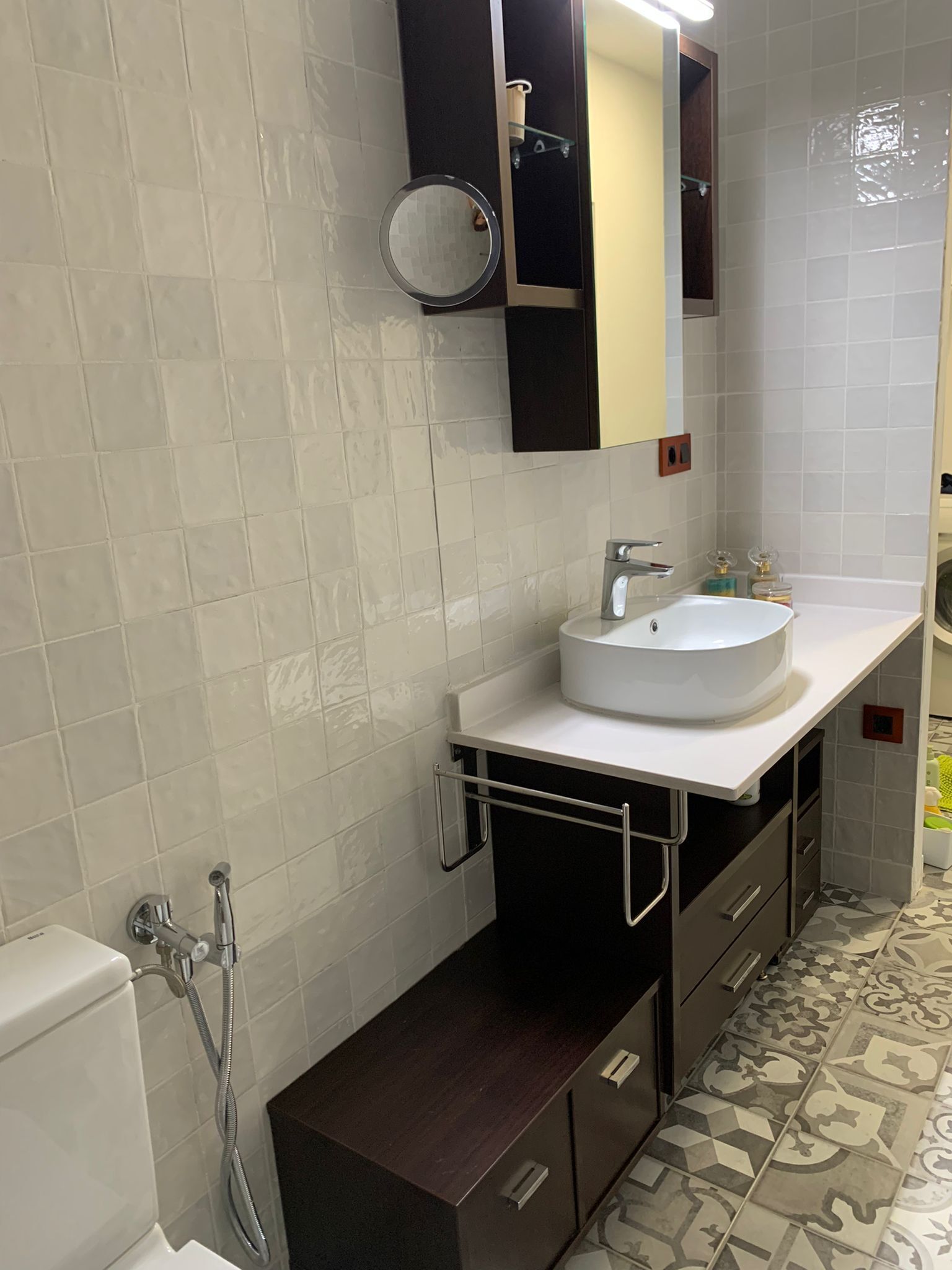 Un baño con lavamanos blanco, mueble de madera oscura y baldosas de piso estampadas.