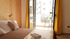 A hotel room with a bed, a window with yellow curtains, and a view of a balcony and building outside. - Independent apartments/rooms  (Otranto)