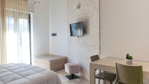 A modern hotel room with a bed, a small seating area, a desk with a chair, and a television mounted on a textured wall. - Independent apartments/rooms  (Otranto)