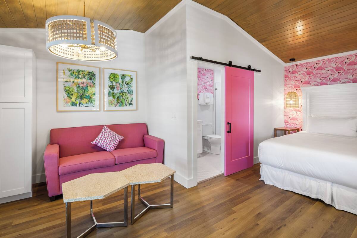 Une pièce avec un canapé rose, un lit, un plafond en bois et une porte blanche donnant sur une salle de bain.