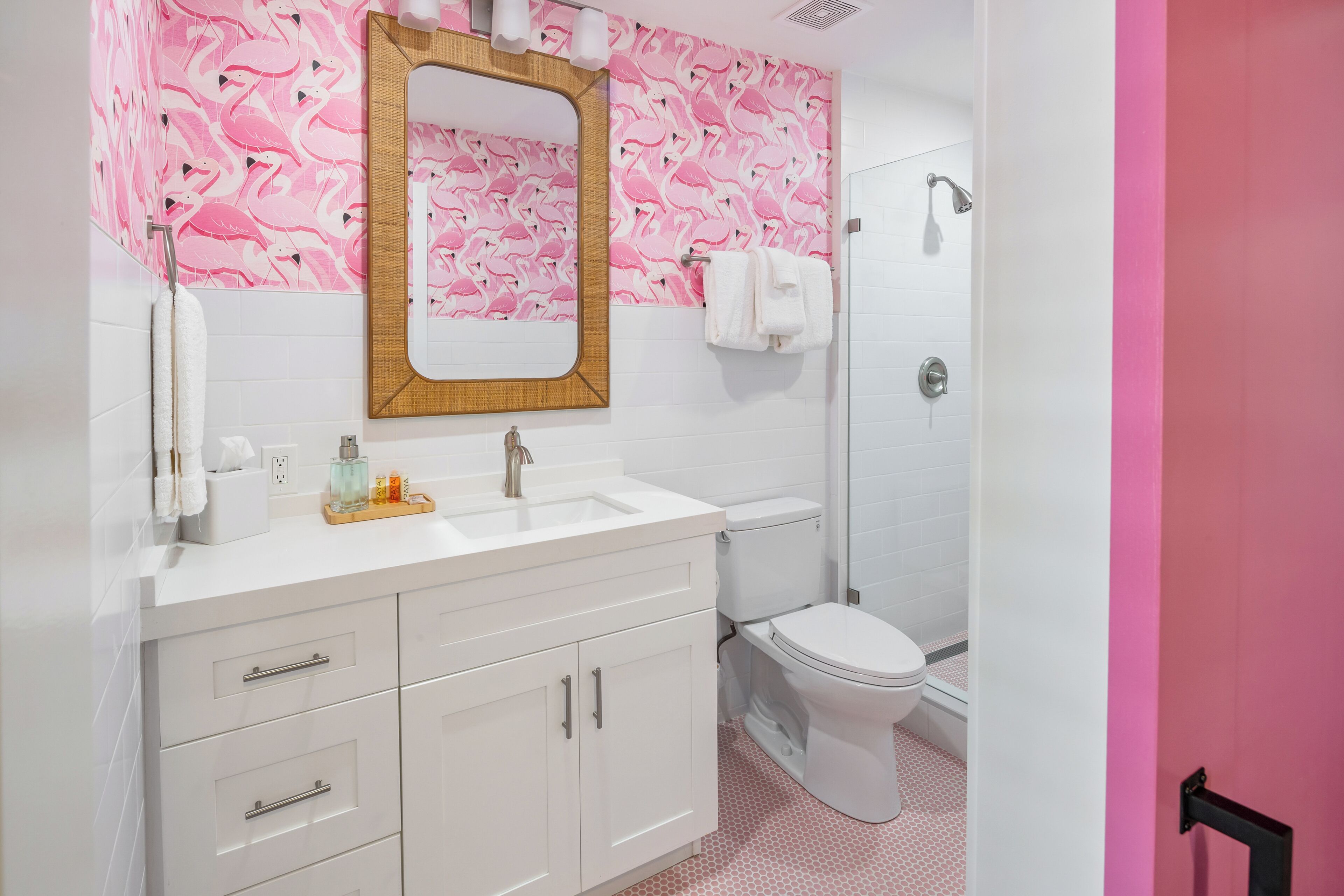 Un baño con lavamanos, espejo y váter blancos, papel tapiz con estampados de flamencos rosas, y una zona de ducha.