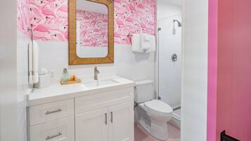 Un baño con lavamanos, espejo y váter blancos, papel tapiz con estampados de flamencos rosas, y una zona de ducha.