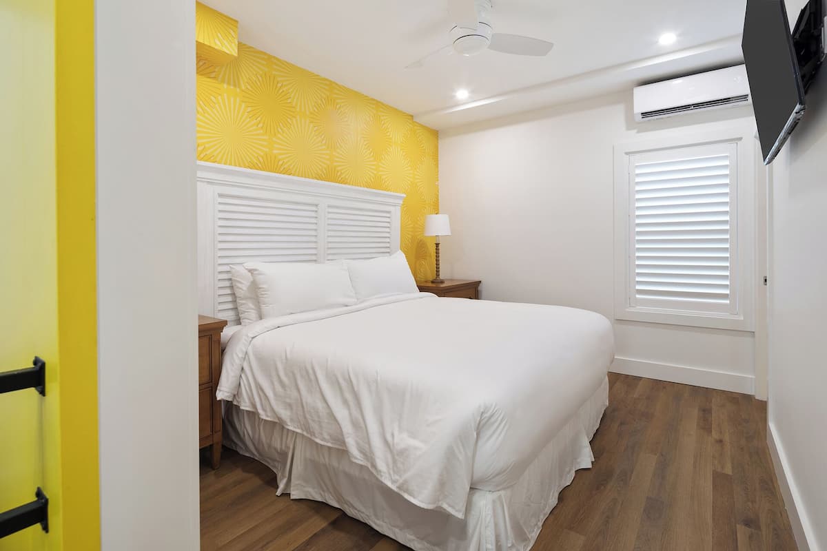 Une chambre à coucher avec un lit, un plancher en bois, des murs blancs, un papier peint à motifs jaunes, un ventilateur de plafond et un appareil de climatisation.
