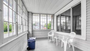 Eine überdachte Veranda mit weißen Stühlen und einem Tisch, großen Fenstern und einer blauen Mülltonne.