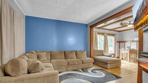 Ein Wohnzimmer mit einem großen beigefarbenen Ecksofa, einem kleineren Sofa, einem Teppich mit Wirbelmuster und einem Essbereich im Hintergrund.