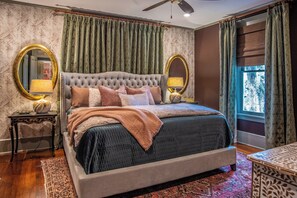 Ein Schlafzimmer mit einer gebügelten Kopfteil, zwei Nachttischlampen, einem großen Spiegel und Blick ins Freie durch ein Fenster mit gemusterten Vorhängen.