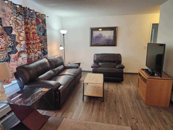 A living room with a leather sofa, a wooden coffee table, a TV on a wooden stand, a floor lamp, and a framed picture on the wall.