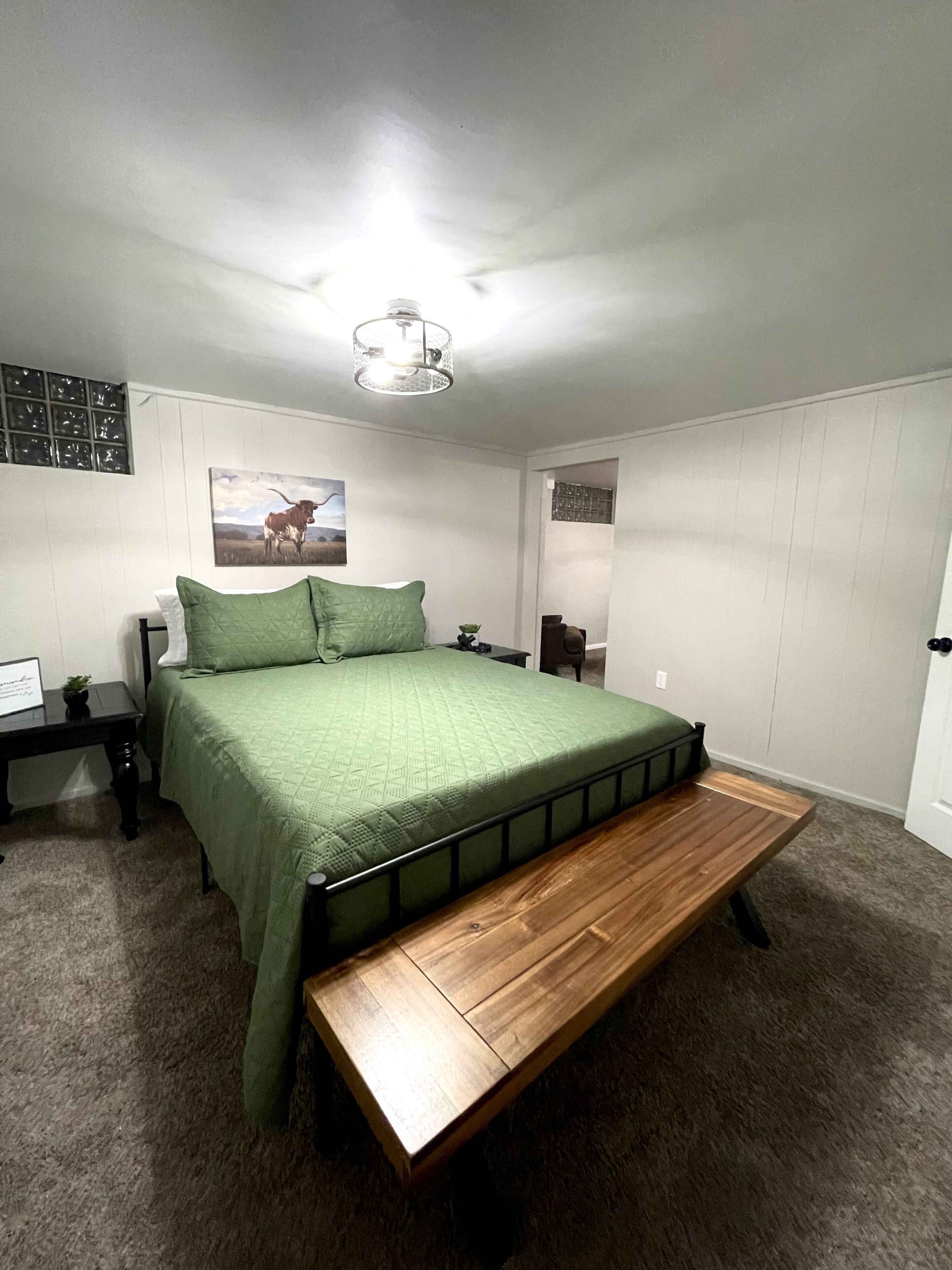 A bedroom with a bed, bedside tables, a nightstand, a wall-mounted light fixture, and a framed picture on the wall.
