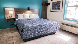 Quarto de hotel com cama, mesa de cabeceira, luminária na parede, obra de arte emoldurada e janela com persianas.