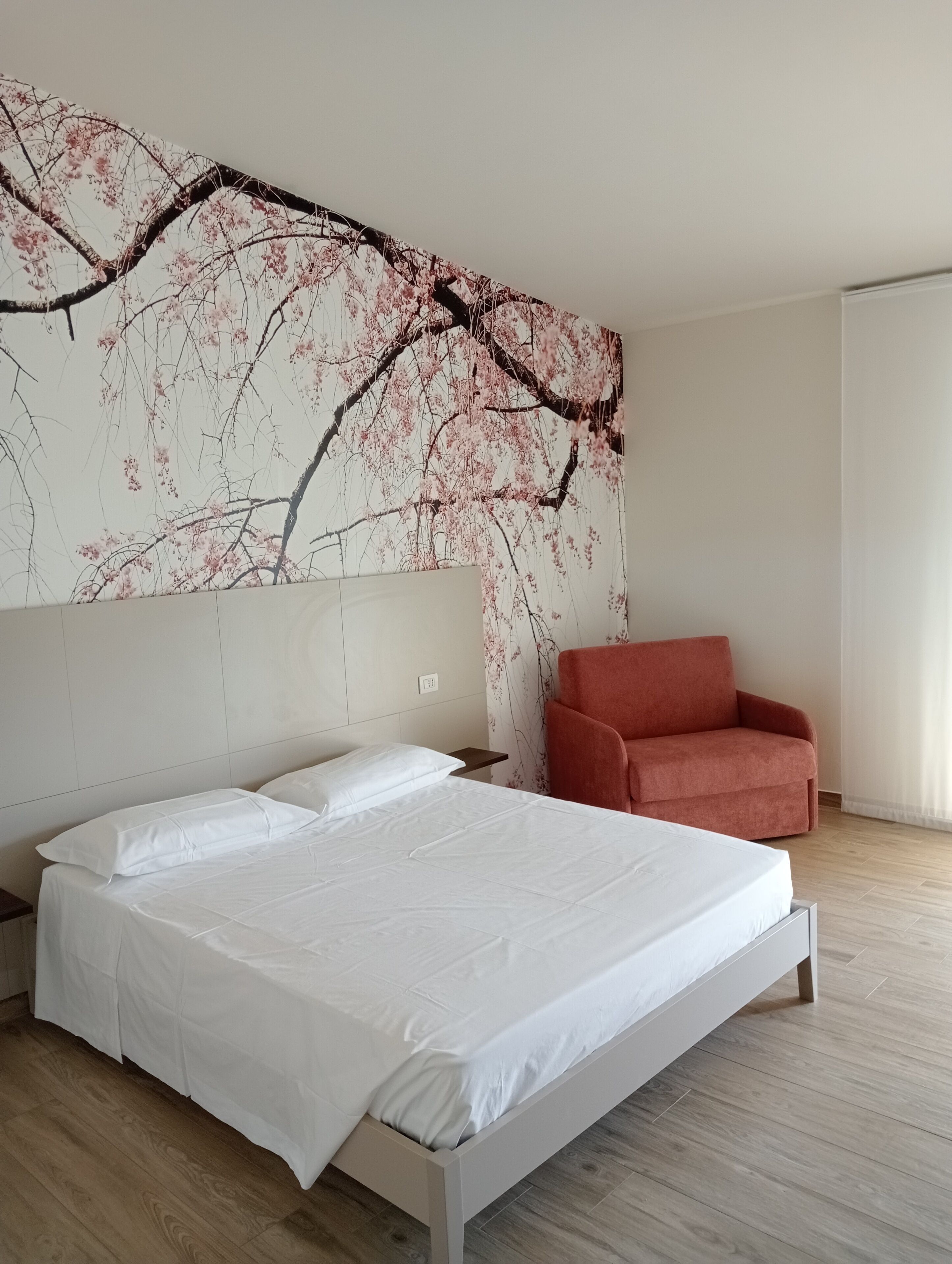 A bedroom with a bed, a nightstand, a red chair, and a wall with a cherry blossom tree mural.