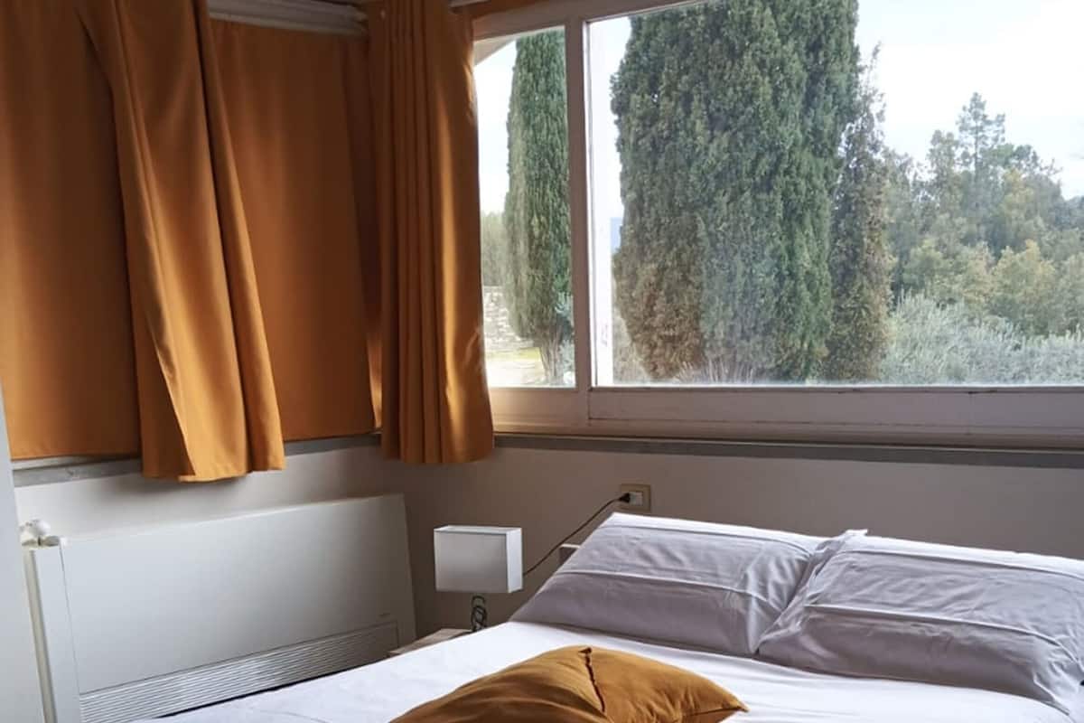 A modern bedroom with a curved bed, white linens, a single mustard-colored pillow, and a window with curtains.
