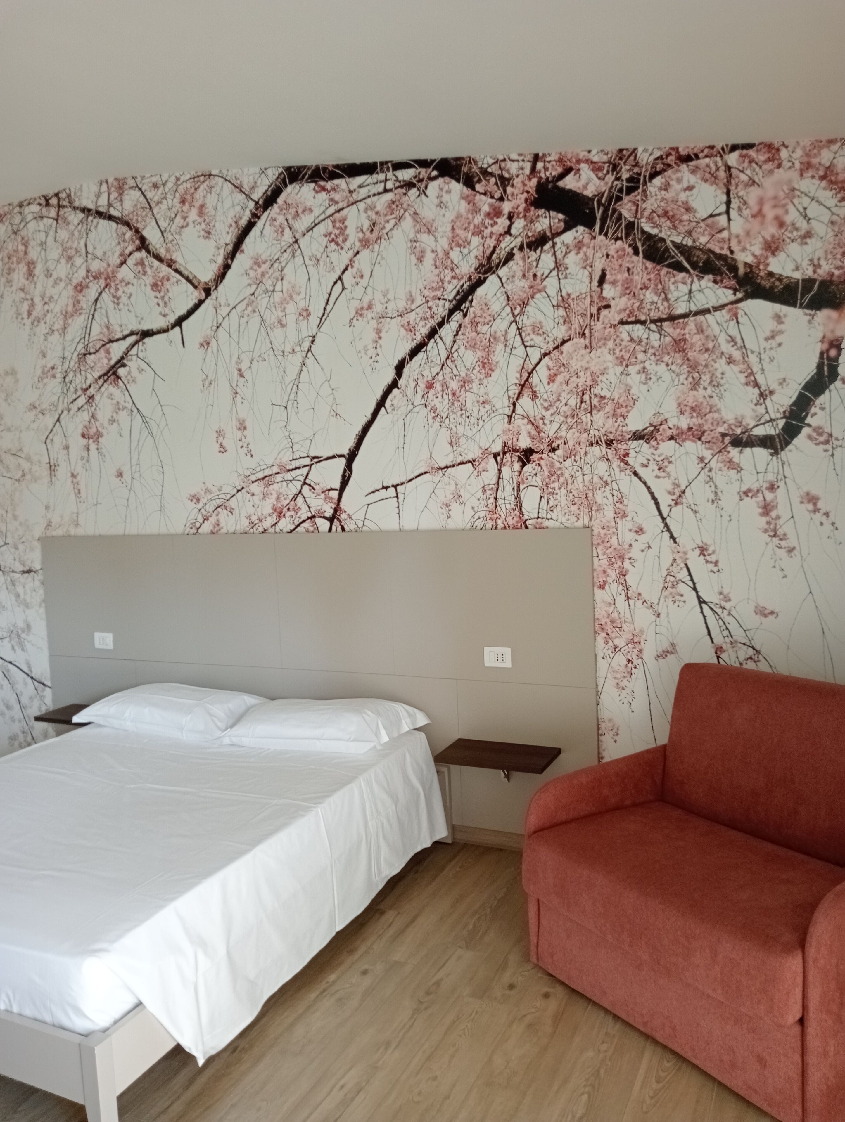 A bedroom with a bed, a nightstand, a red sofa, and a wall with a cherry blossom mural.
