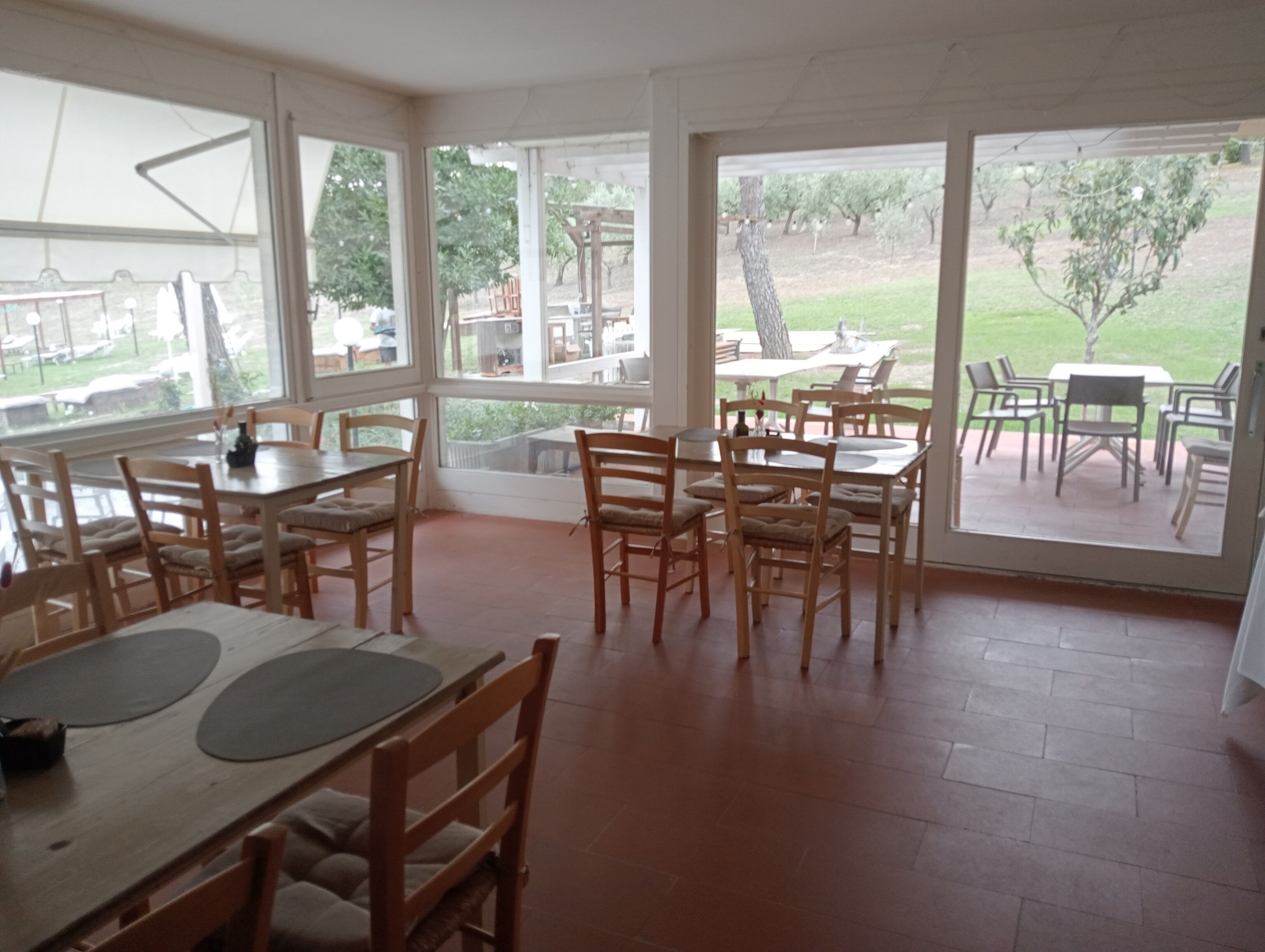 An indoor dining area with wooden tables and chairs, large windows, and a view of an outdoor patio.