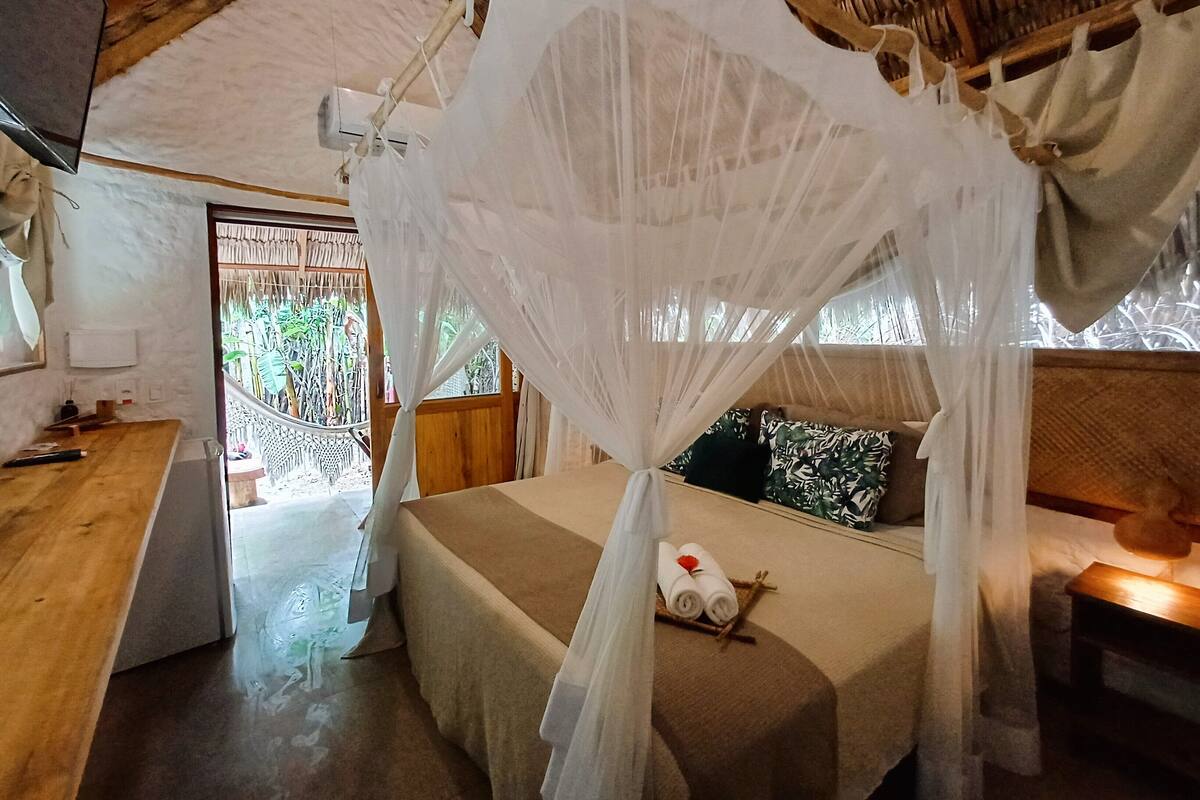 Quarto com cama sob um dossel branco, uma escrivaninha de madeira e janela com vista para o verde.