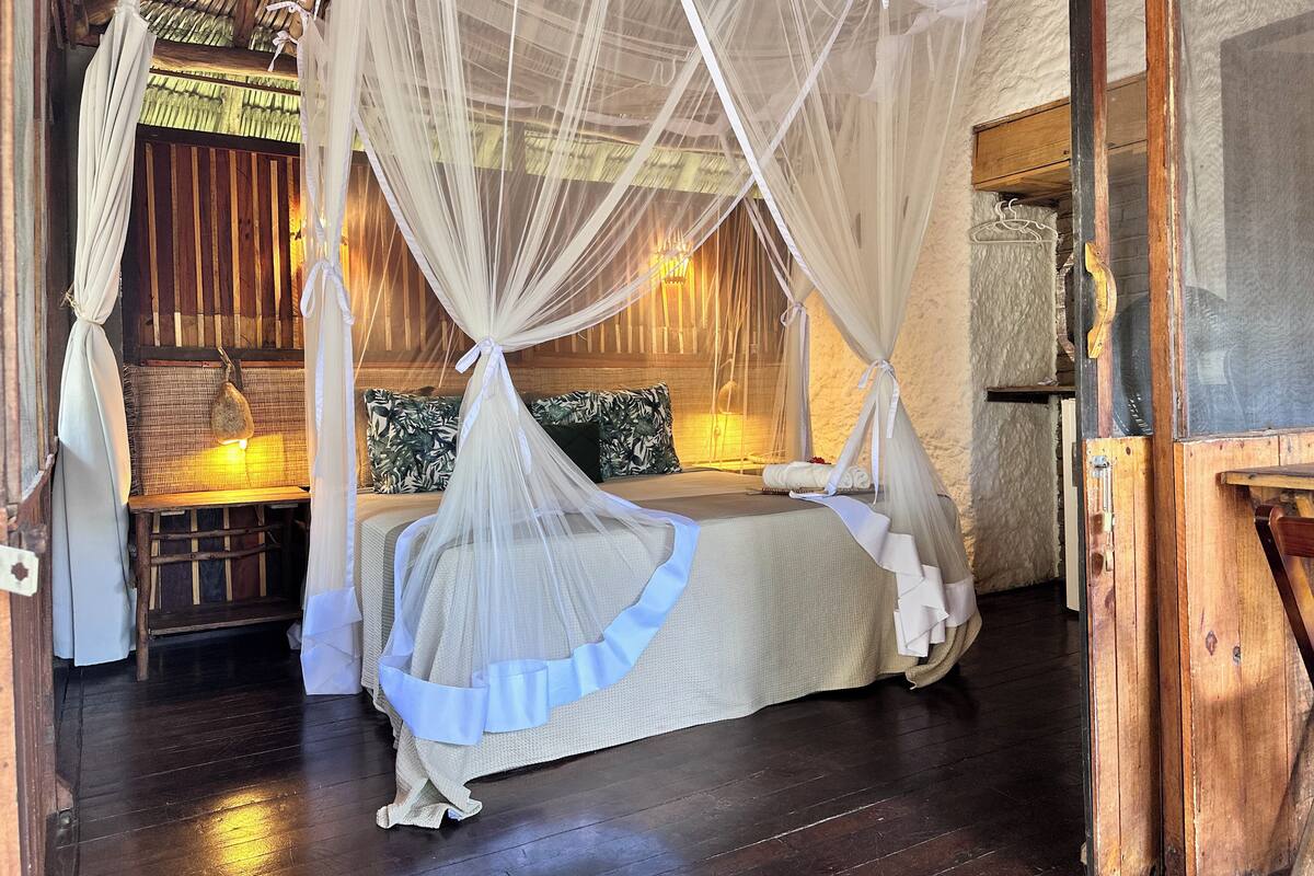 Quarto com cama sob dossel, um banco de madeira e um banco de vime com almofadas.