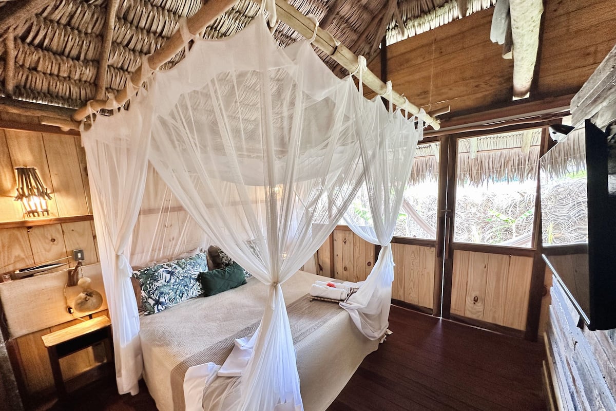 Quarto com cama sob um dossel branco, paredes de madeira e janela com vista para o exterior.