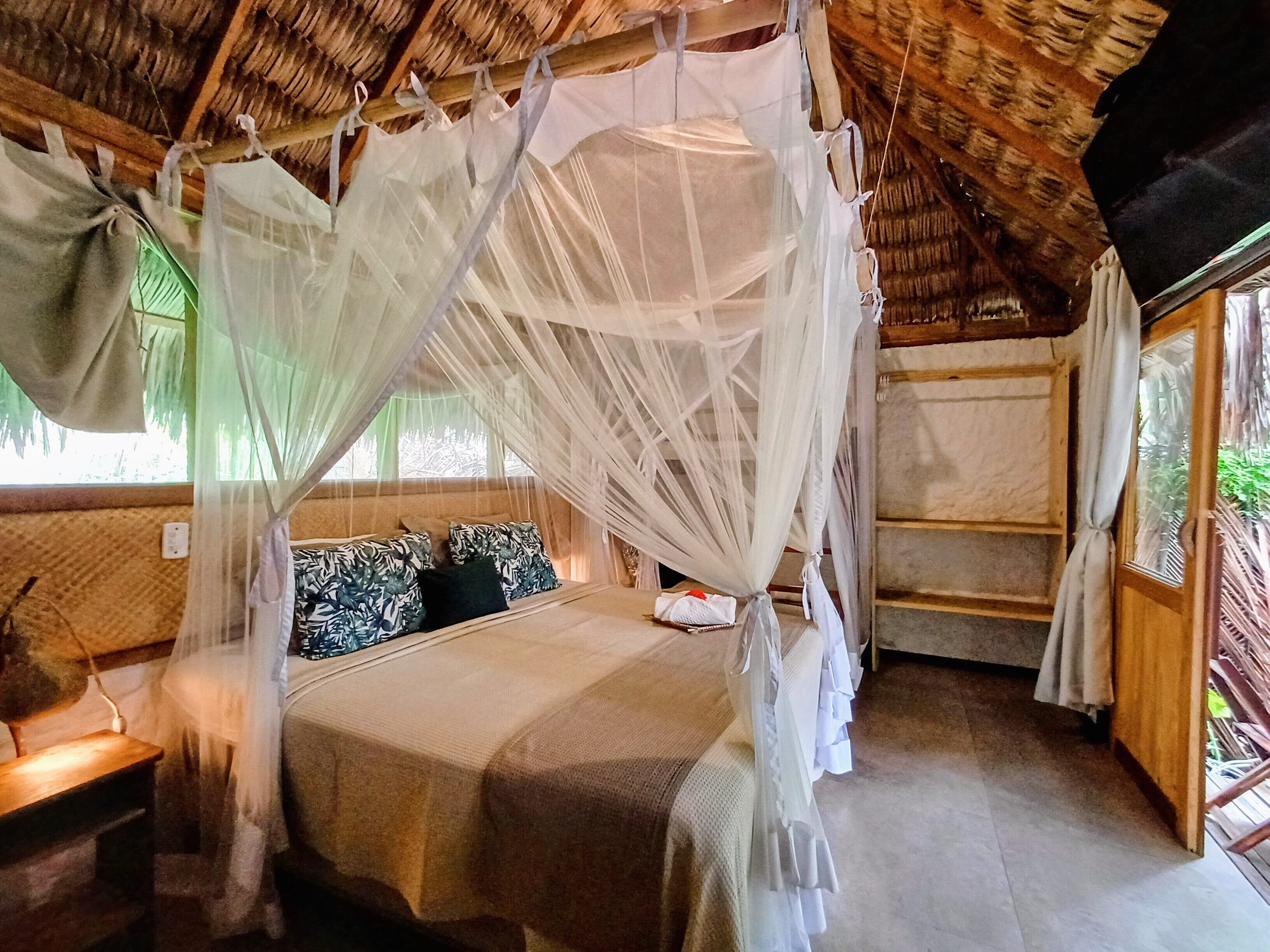 A room with a bed under a mosquito net, wooden walls, and a window with curtains.