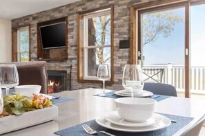 A dining area with a stone fireplace, a flat-screen TV mounted on the wall, and a view of the outdoors through large windows.