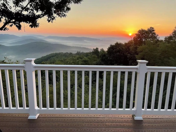A wooden deck with white railings overlooking a mountainous landscape during sunset.