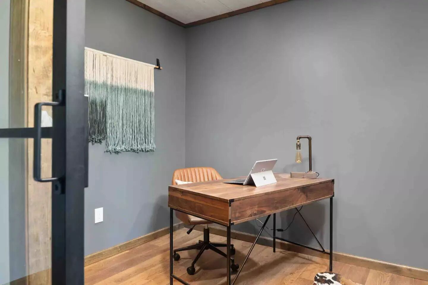 A room with a wooden desk, a laptop, a chair, a lamp, and a wall hanging.